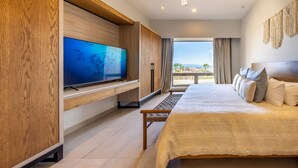 A modern hotel room with a large flat-screen TV mounted on a wooden wall, a large bed with multiple pillows, a ceiling fan, and a balcony with a view of the mountains.