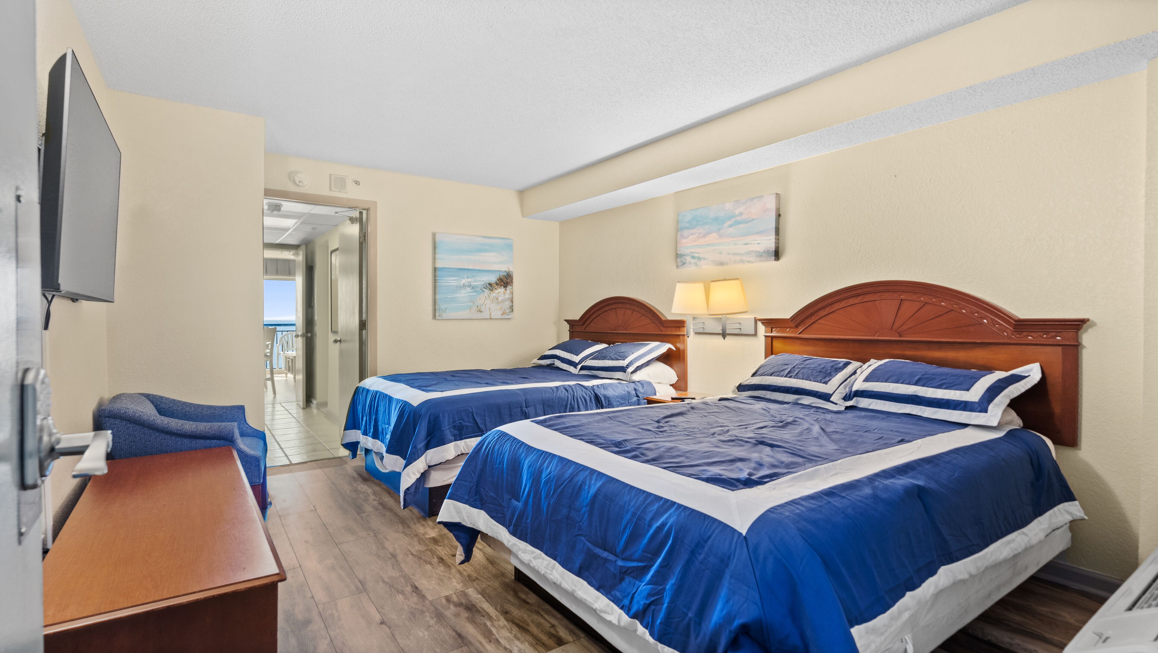 A hotel room with two beds, a TV, a desk, and a view of the beach.