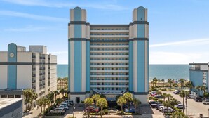A multi-story hotel with a central entrance, parking lot, and a view of the ocean. - Direct oceanfront, separate bedroom, full kitchen - rates lower than last year ! (Myrtle Beach)
