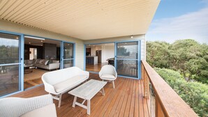 A spacious balcony with wooden flooring, white wicker furniture, and large glass doors leading to an interior living area.