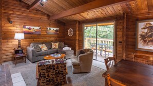 L’intérieur d’un chalet confortable, avec des murs en rondins, comprend un canapé, un fauteuil et une table à manger. Des tableaux ornent les murs et une porte-fenêtre coulissante offre une vue sur l’extérieur.