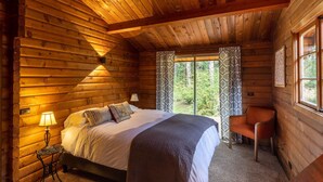 Une chambre dans un chalet en rondins, avec un lit, une chaise, une lampe et une fenêtre avec des rideaux.