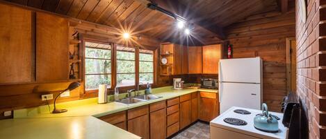 A rustic cabin kitchen with wooden walls, a large window, a sink, a stove, and a refrigerator.