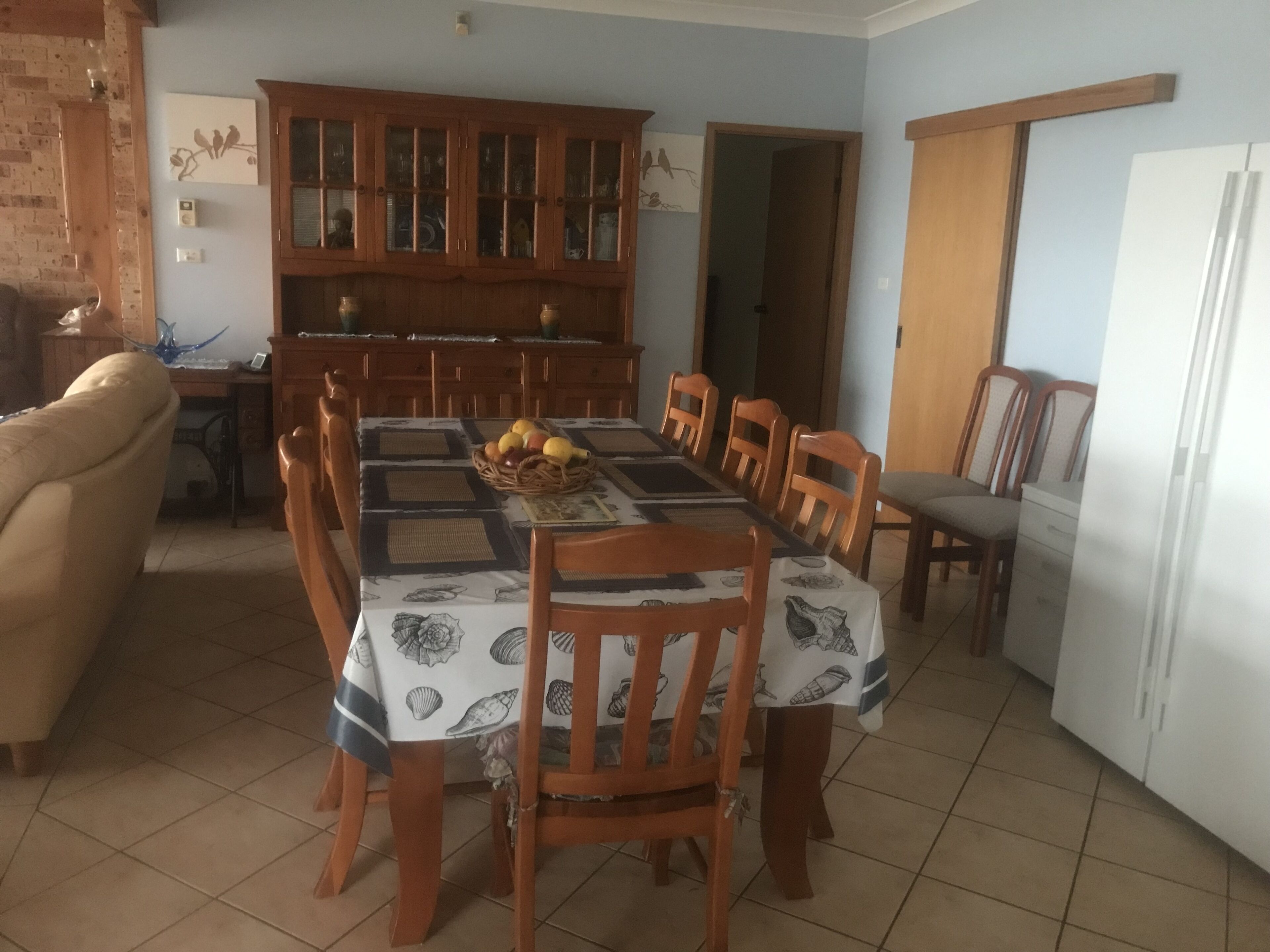 Zona de menjador amb una taula preparada per a sis persones, envoltada de cadires de fusta. Darrere de la taula hi ha un armari de fusta amb portes de vidre, i una nevera a la dreta.