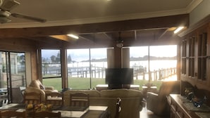 Amplia sala de estar con televisor grande, asientos cómodos, zona de comedor con mesa preparada para cuatro personas y vista a un embarcadero y al agua.