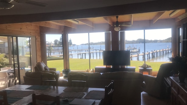 Amplia habitación con grandes ventanas que dan a un muelle y al agua. Hay un televisor sobre un mueble bajo y una mesa de comedor con sillas.