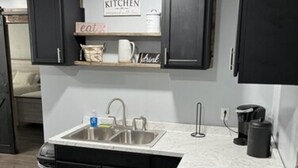 Eine moderne Küche mit schwarzen Schränken, einer weißen Arbeitsplatte und einem Edelstahlspülbecken. Über dem Spülbecken ist ein Schild mit der Aufschrift 'KITCHEN' angebracht, und ein weiteres Regal enthält Artikel mit den Aufschriften 'eat' und 'drink'.