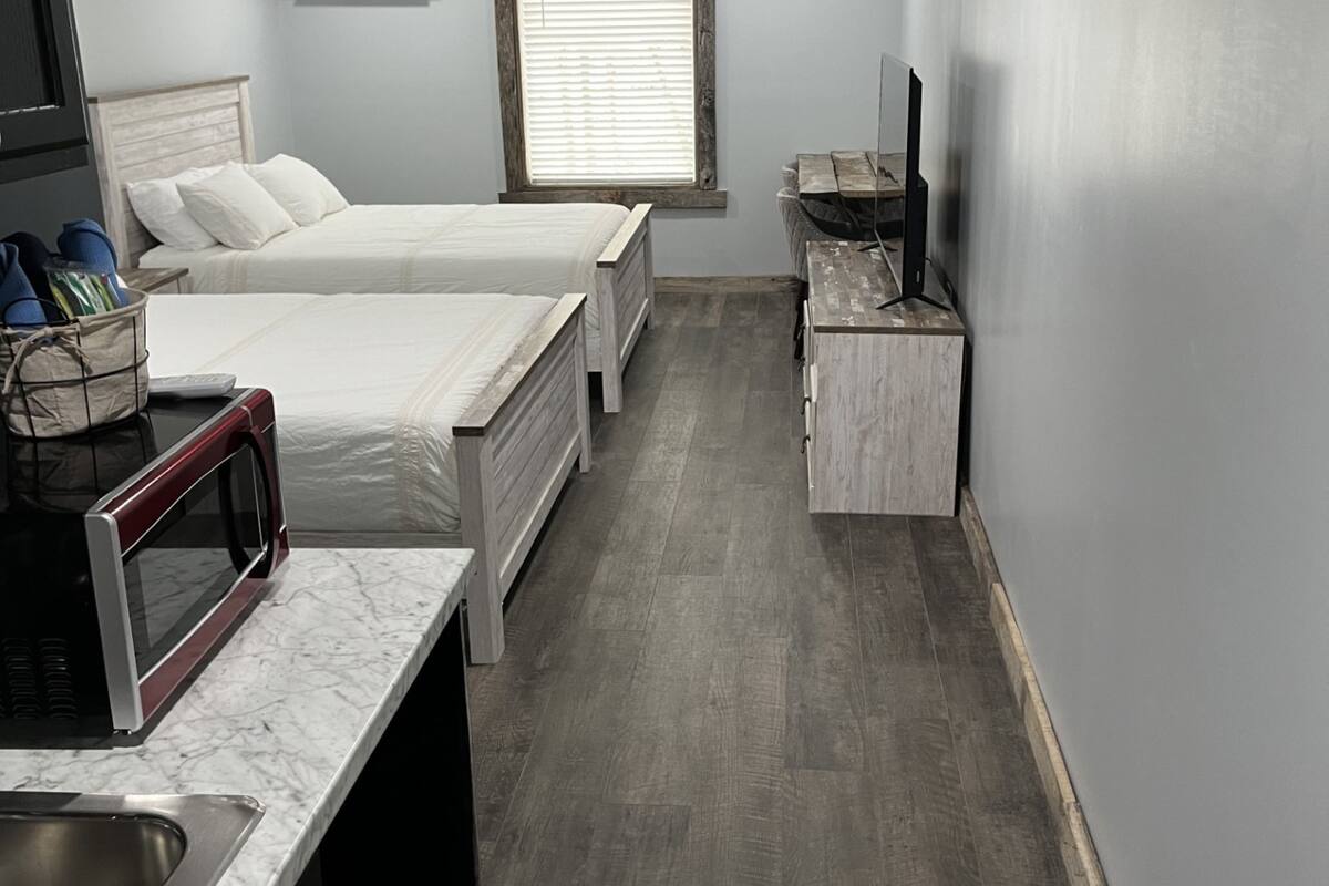 A compact hotel room with two beds, a small kitchenette, a TV, and a window with blinds.