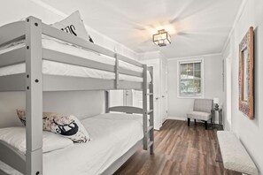 A bunk bed room with a wooden floor, a chair, a bedside table, and a window with blinds.