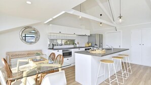 Een moderne keuken met witte kastjes, een centraal eiland met barkrukken, en een eetgedeelte met een glazen tafel en stoelen.