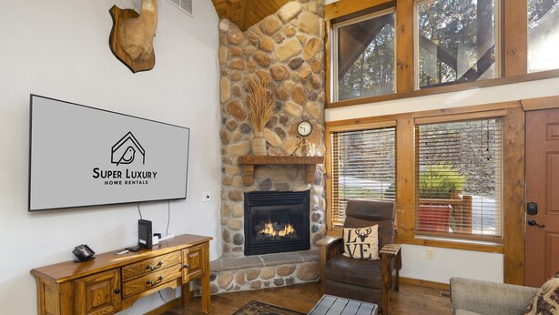 A cozy living room with a stone fireplace, wooden furniture, and a large window with blinds.