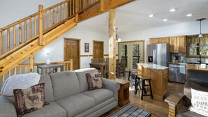 A spacious living area with a wooden staircase, a gray sofa with decorative pillows, a dining table, and a kitchen area with wooden cabinets and stainless steel appliances.