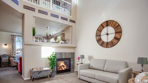 A spacious living room with a fireplace, a large wall clock, and a high ceiling with a balcony.