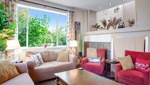 A living room with a beige sofa, a red sofa, and a wooden coffee table. There is a window with curtains, a fireplace with decorative items, and a plant on the windowsill.
