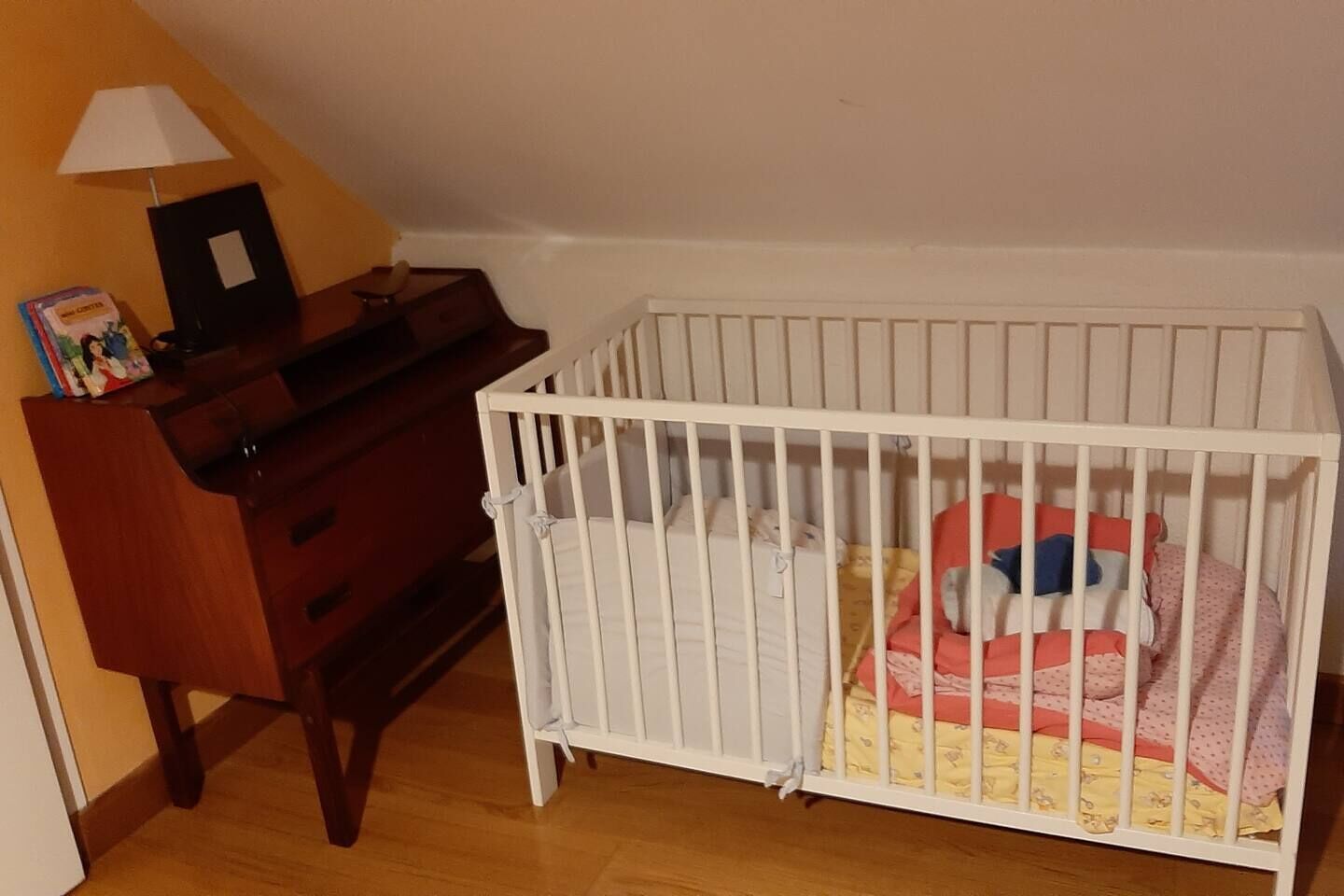 A room with a white crib, wooden dresser, and a lamp.