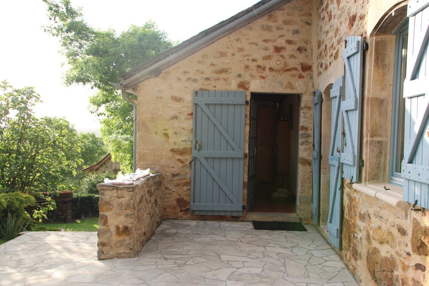 A rustic stone building with a wooden door and shutters, a stone bench, and a paved patio.