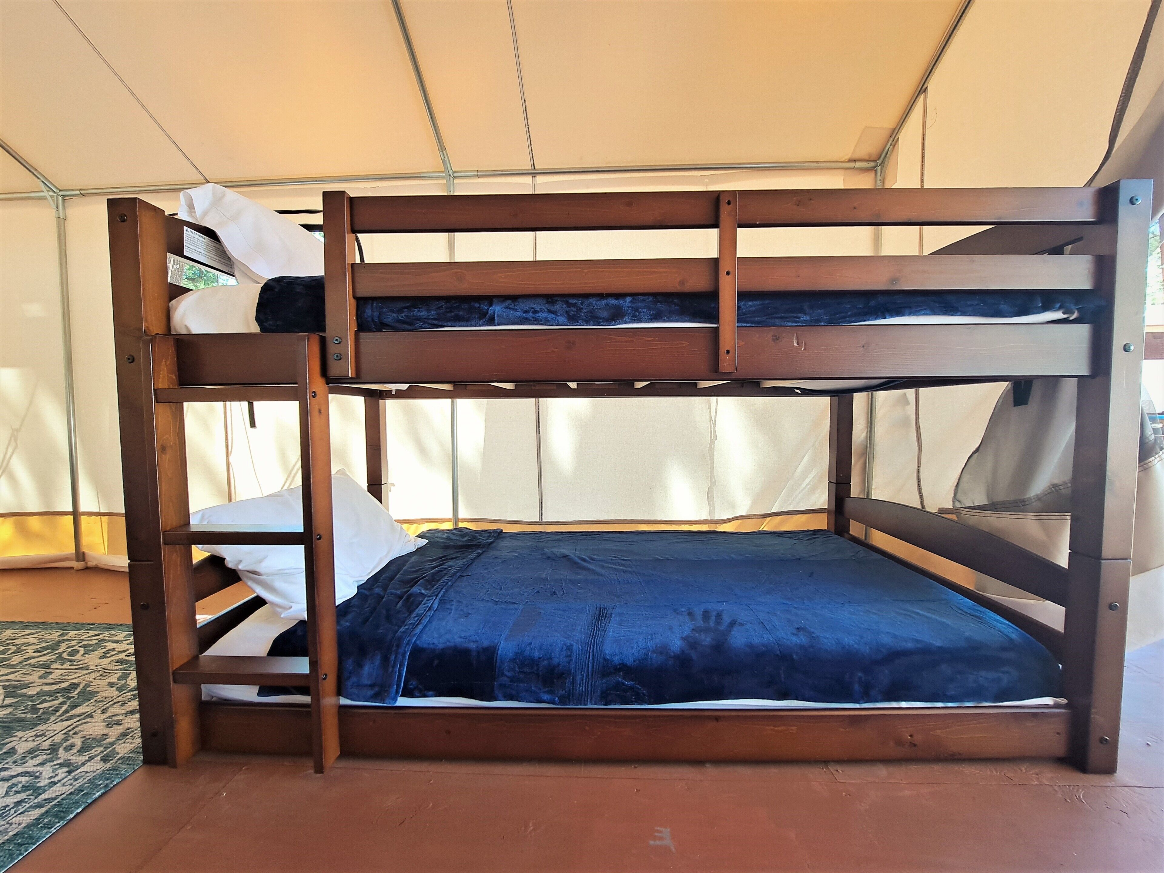 A bunk bed with a blue mattress and white pillows, set in a tent-like structure with a green patterned rug on the floor.