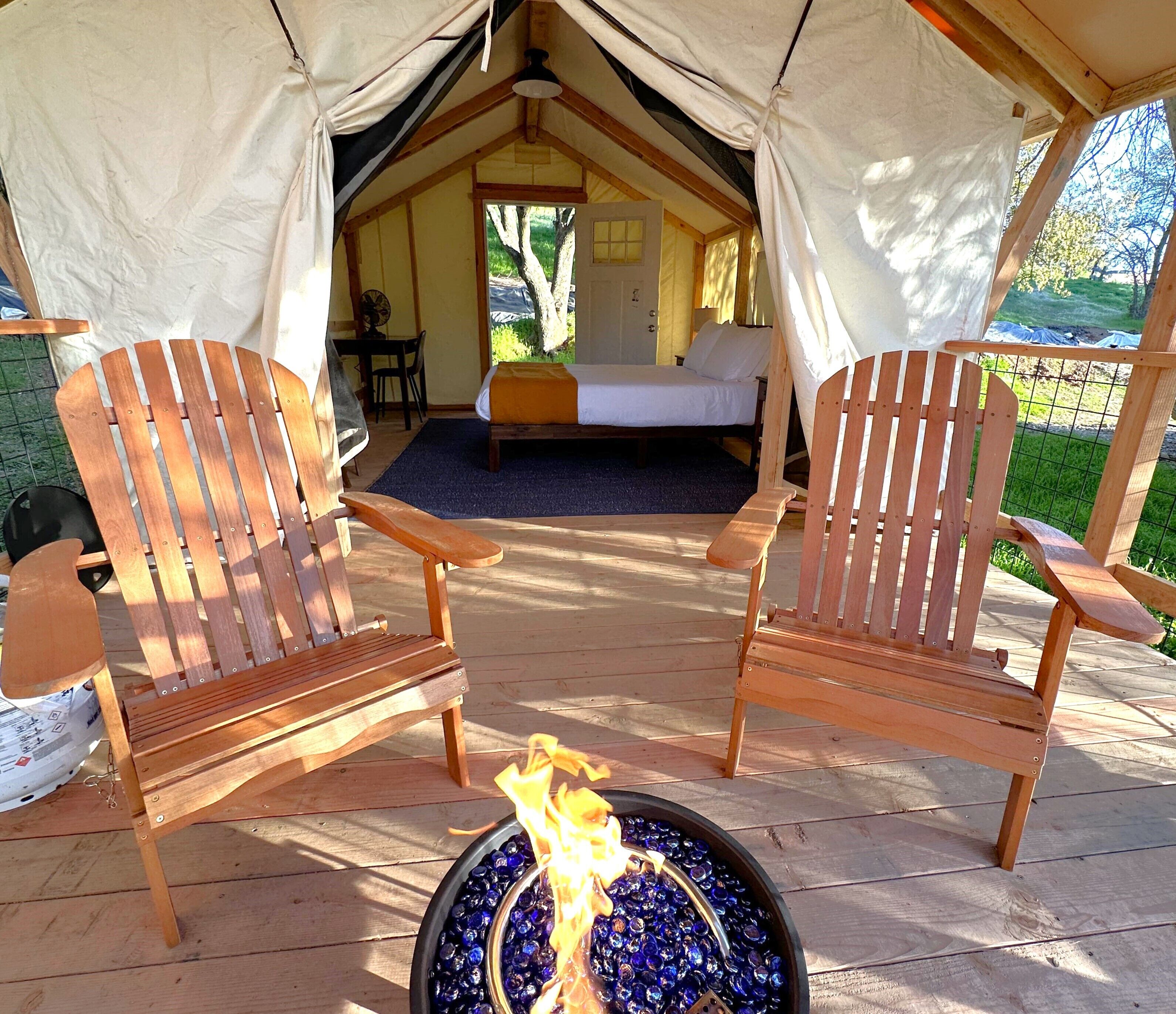 A tented accommodation with two wooden chairs, a bed, and a fire pit with blue glass bottles.