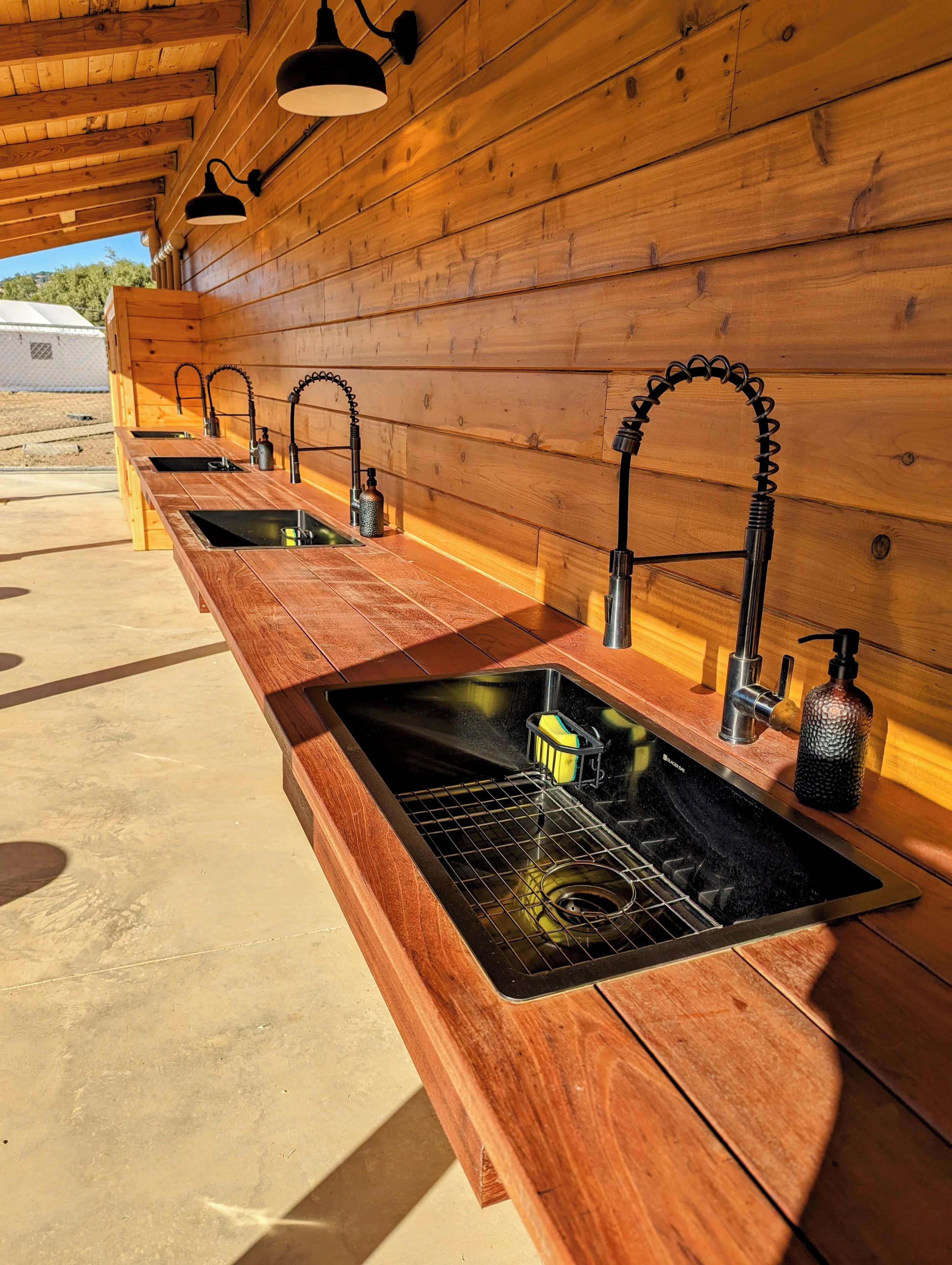 A row of wooden sinks with modern faucets and soap dispensers, set in a sunny outdoor area with wooden walls.