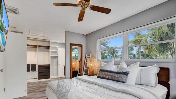 Un dormitorio con una cama grande, ventilador de techo, ventana con vista a palmeras y vestidor.
