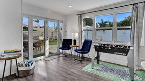 Una habitación con mesa de fútbol, dos sillas azules, una mesita con toallas y vistas al exterior a través de amplias ventanas.