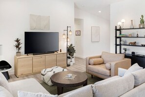 Ein modernes Wohnzimmer mit einem Flachbildfernseher, einem beigefarbenen Sofa, einem Couchtisch und einem Bücherregal mit Dekorationselementen.