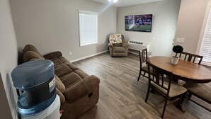 Ein Wohnzimmer mit einem braunen Ecksofa, einem Esstisch mit Stühlen, einem Deckenventilator, einem an der Wand montierten Fernseher und einer kleinen Topfpflanze.