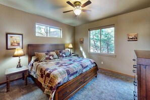 Une chambre dotée d’un grand lit, de deux tables de chevet avec des lampes, d’un ventilateur de plafond et de deux fenêtres donnant sur la verdure.