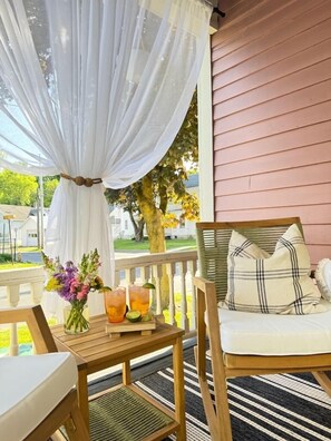 A wooden porch with a cushioned chair and a table, a white canopy, and a vase of flowers.