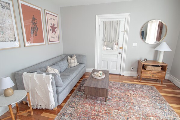 A living room with a grey sofa, a coffee table, a patterned rug, a small side table with a lamp, and a wooden cabinet.