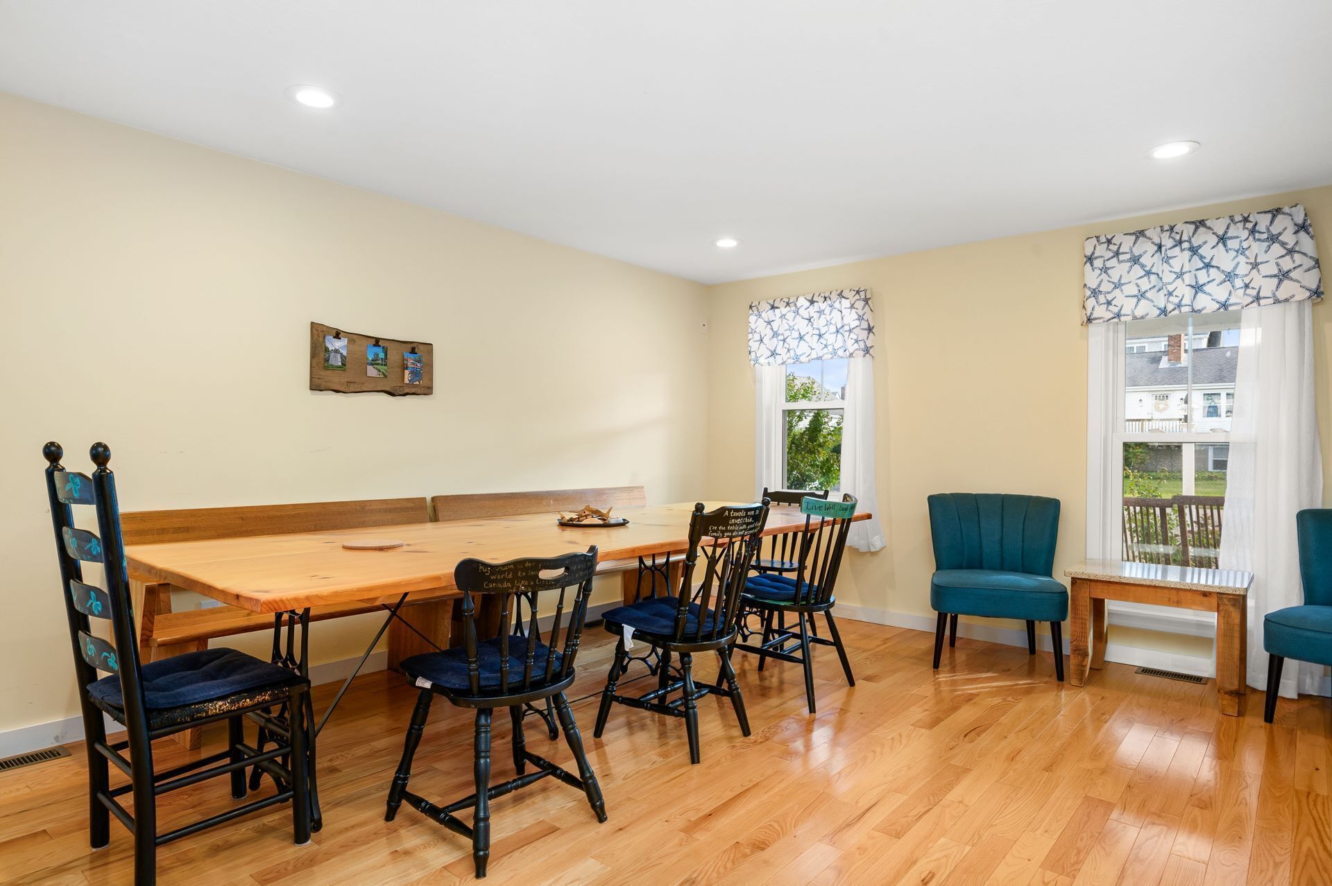 A dining area with a wooden table and chairs, a bulletin board with photos, and windows with curtains.