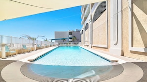 Una piscina con lettini e un ombrellone, circondata da un muretto e un edificio con balconi.