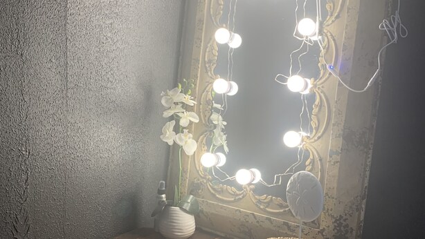Une coiffeuse avec un miroir, entourée de lampes fixées au mur, d’une petite plante en pot et d’un panier décoratif.
