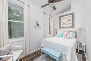 Quarto com cama, ventoinha no teto, janela, mesa de cabeceira e criado-mudo.