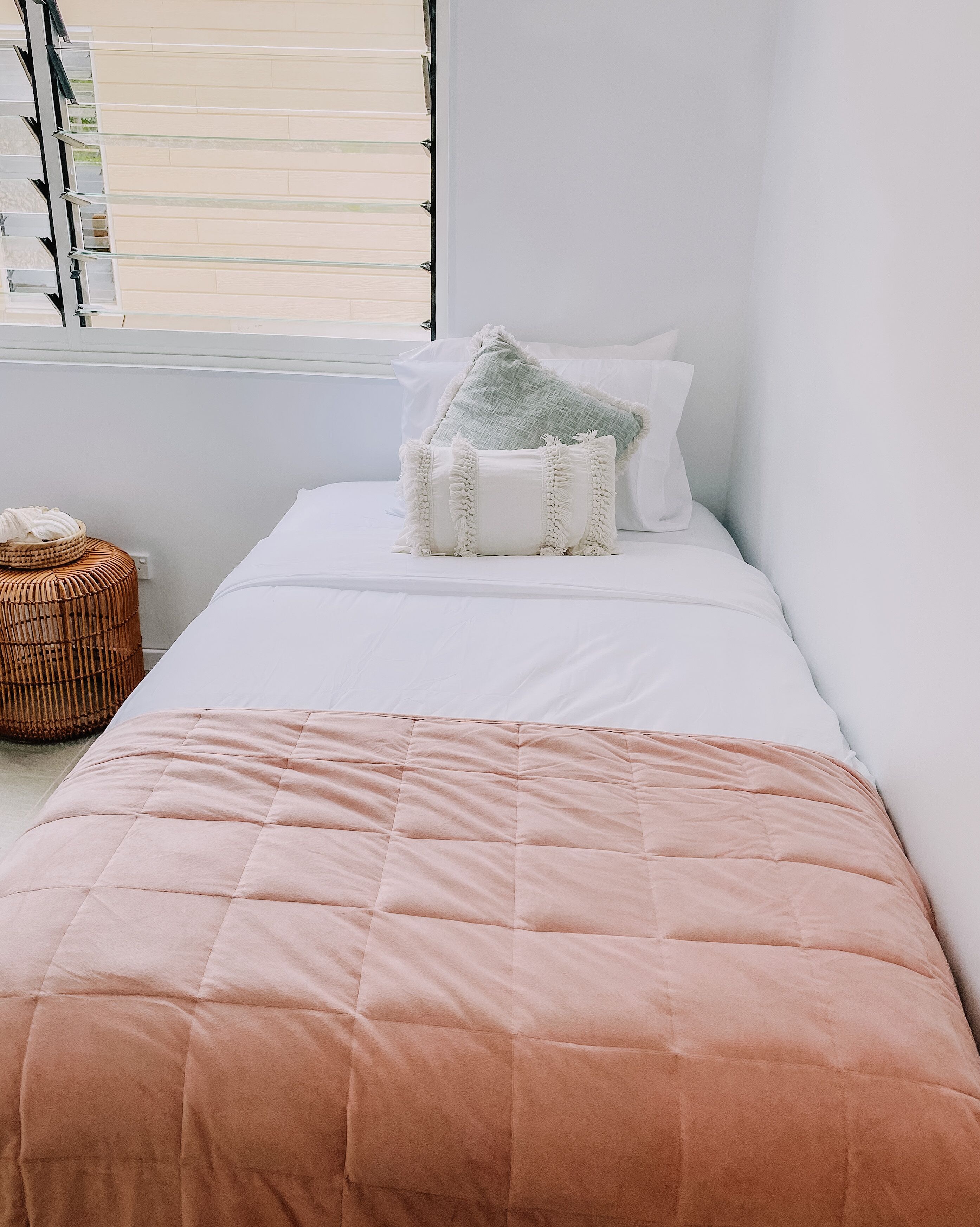 Ein ordentlich gemachtes Bett mit einer rosa Quilterdecke, weißen Kissen und einer grün-weißen Tagesdecke. Auf dem Boden neben dem Bett steht ein Korbwägelchen.