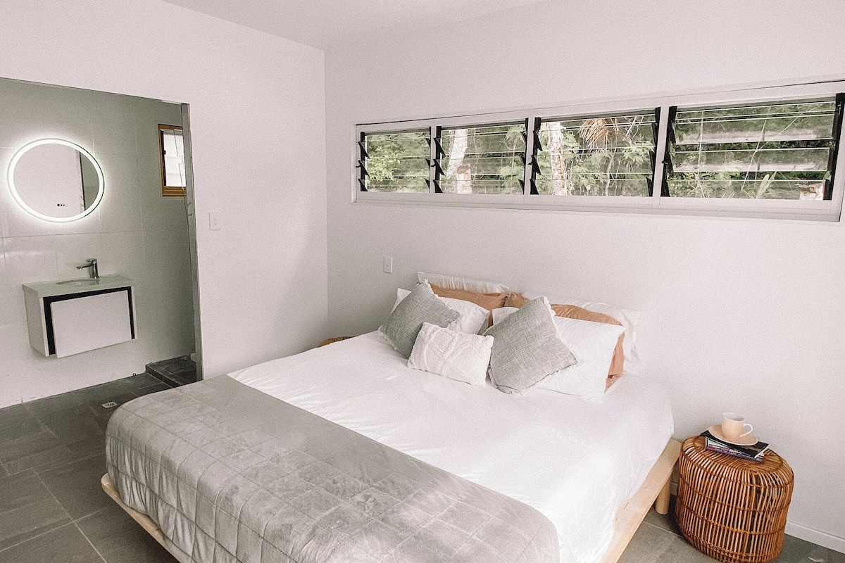 A bedroom with a bed, a bedside table, a mirror, a window with a view, and a bathroom with a sink and mirror.