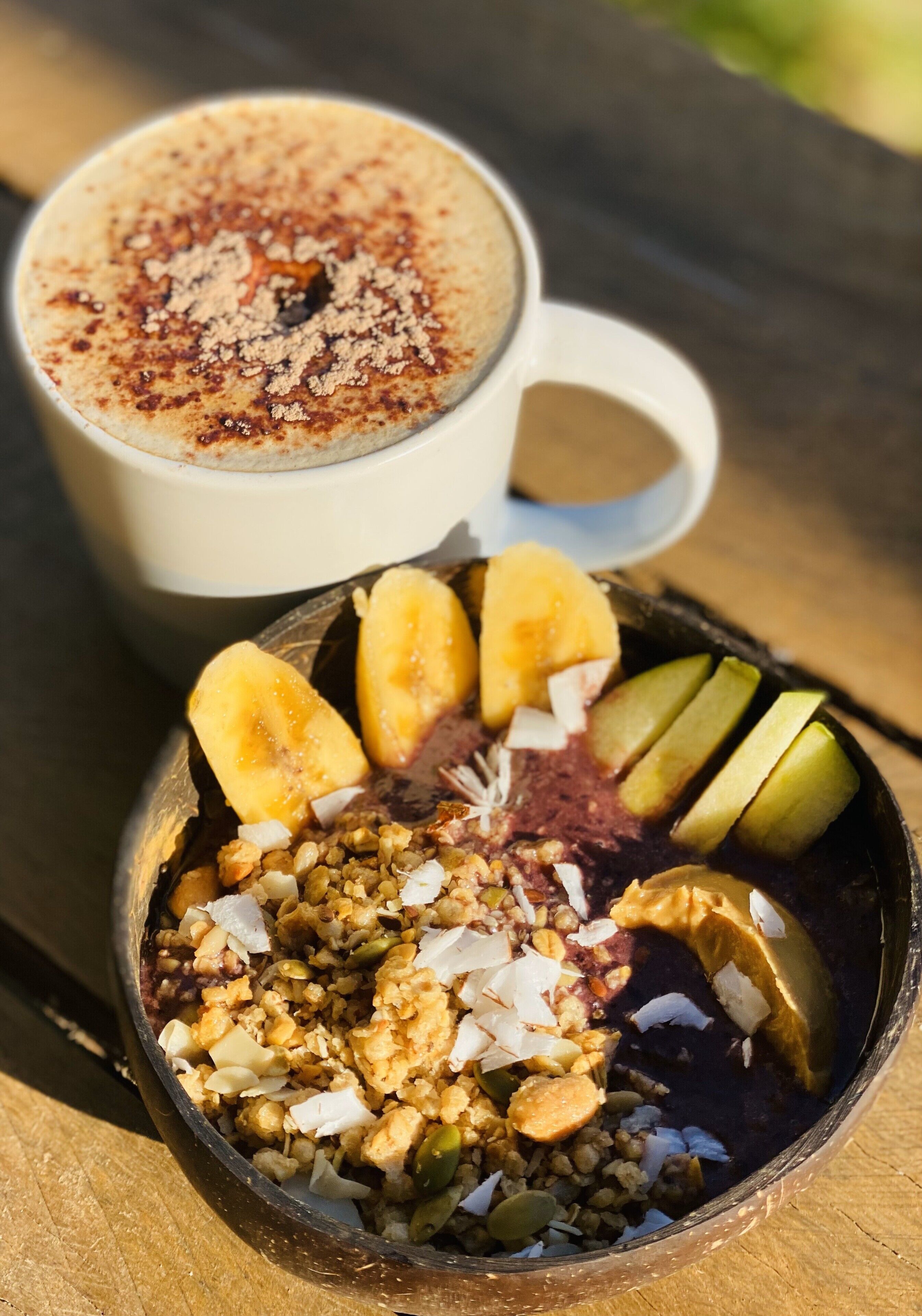 A bowl of granola with banana slices, nuts, and coconut flakes, and a cup of coffee with a frothy top.