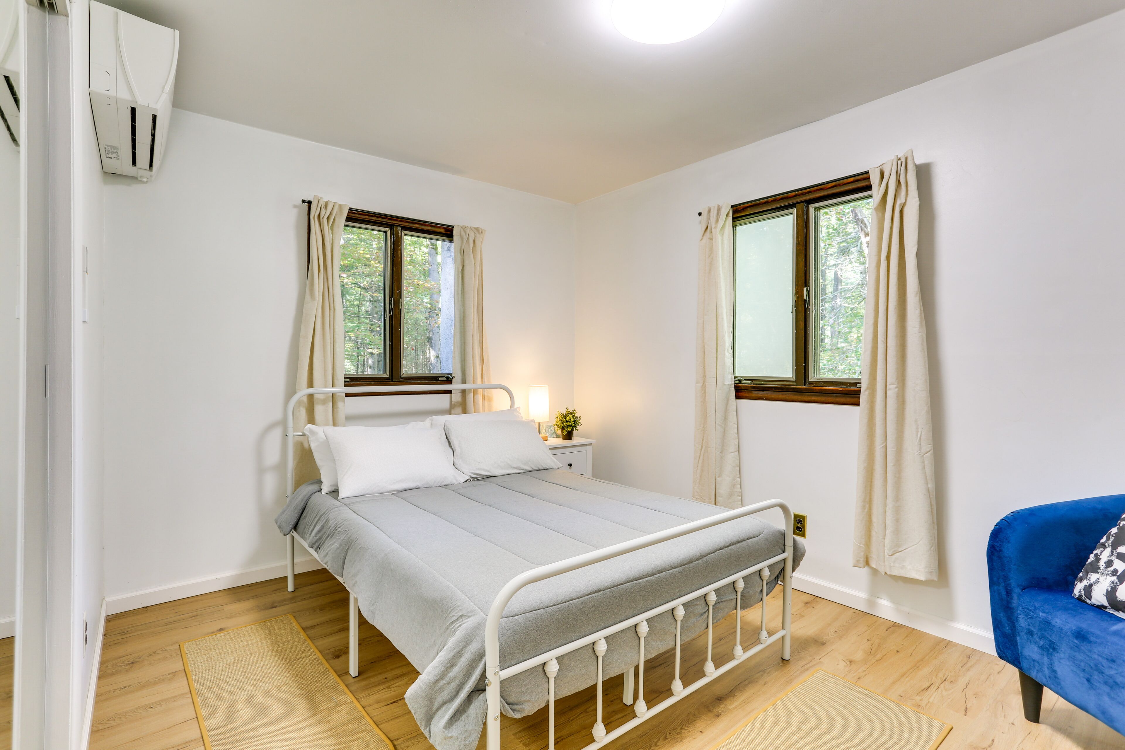 Ein Schlafzimmer mit einem Metallbettgestell, zwei Vorhängen an den Fenstern, einem Nachttisch mit Lampe und einem blauen Sessel.