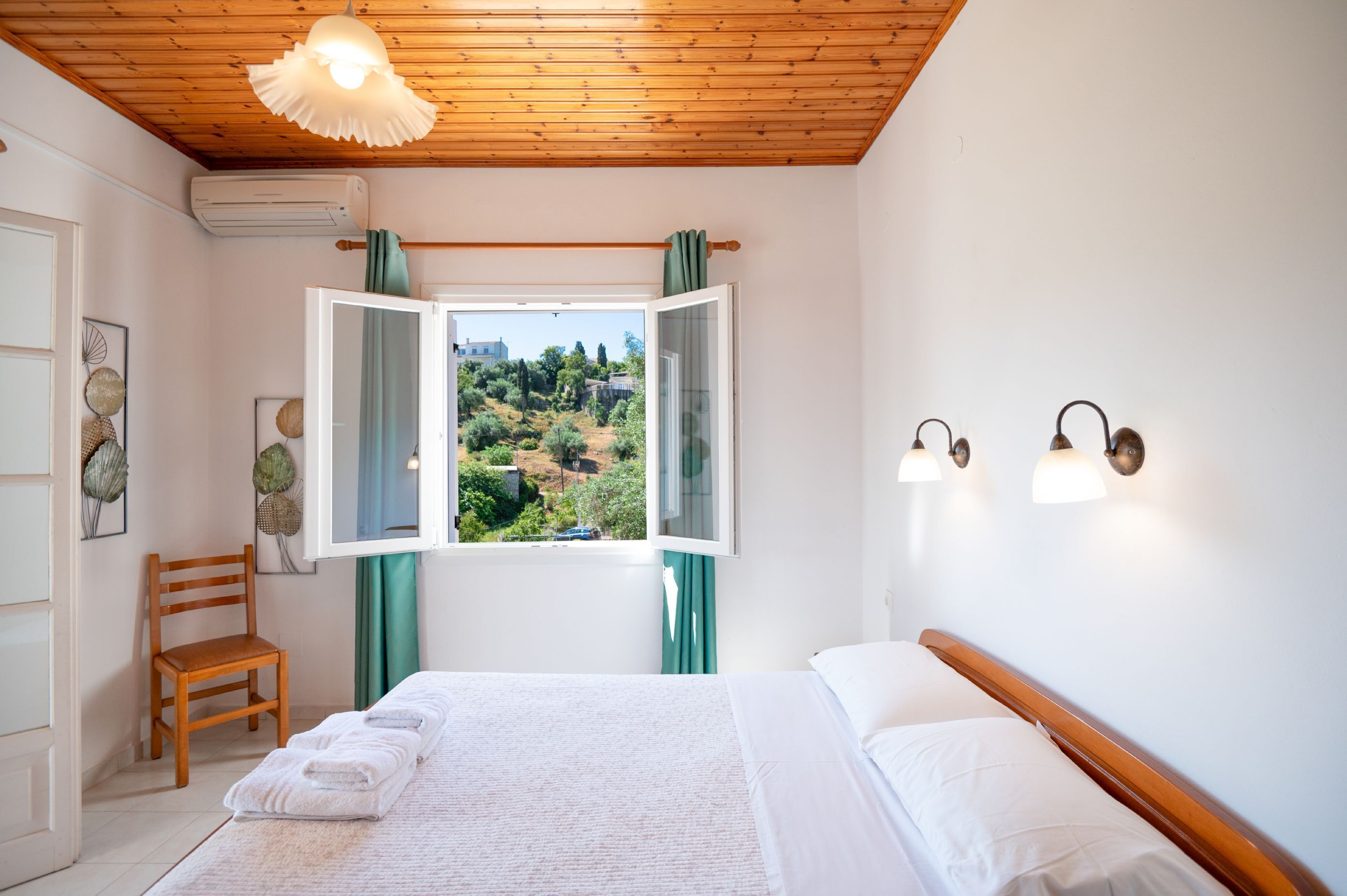 Ein Schlafzimmer mit Holzboden, einem Bett mit weißen Leinen, einem Stuhl und Blick auf eine grüne Landschaft durch ein offenes Fenster.