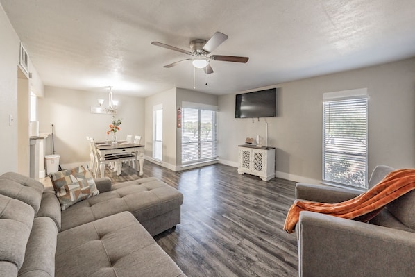 A spacious living room with a large sectional sofa, a dining area with a table and chairs, a ceiling fan, and a flat-screen TV mounted on the wall.