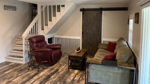 Un salón con un sillón de cuero marrón, un sofá beige con cojines rojos, una mesa de centro de madera y una escalera con barandillas blancas.