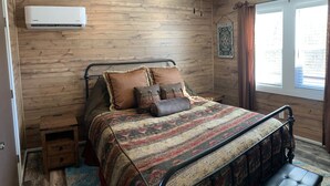 Un dormitorio con pared de madera, una cama con ropa de cama estampada, un ventilador de techo y un aire acondicionado.
