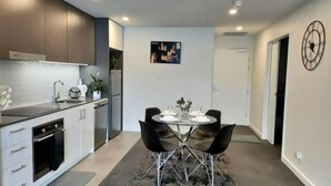 Una cocina moderna con gabinetes blancos, encimera negra y electrodomésticos de acero inoxidable. Hay un comedor con una mesa redonda y sillas negras, y un reloj en la pared.