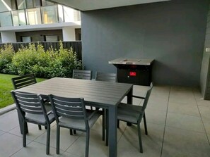 Un patio al aire libre con una mesa rectangular y seis sillas, una parrilla incorporada y vistas a un edificio moderno al fondo.