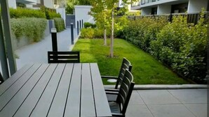Un patio moderno al aire libre con una mesa rectangular y sillas, rodeado de un césped cuidado y setos bajos.