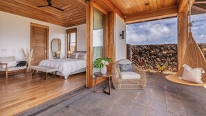A spacious bedroom with a large bed, a sitting area with a wicker chair, and a view of a stone wall and sky.