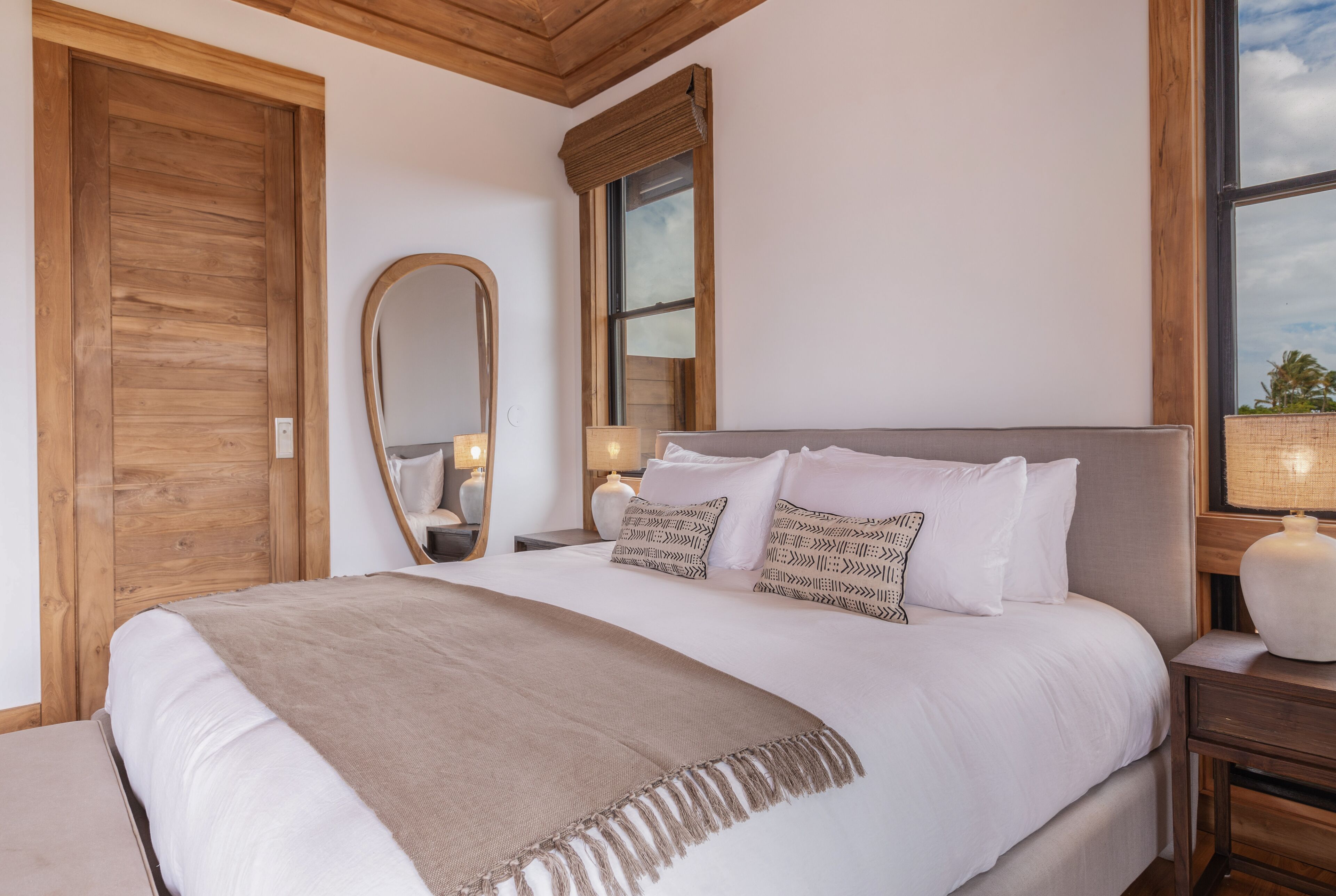 A bedroom with a large bed, wooden headboard, a mirror, bedside lamps, and a window with a view of palm trees.