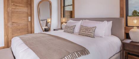 A bedroom with a large bed, wooden headboard, a mirror, bedside lamps, and a window with a view of palm trees.