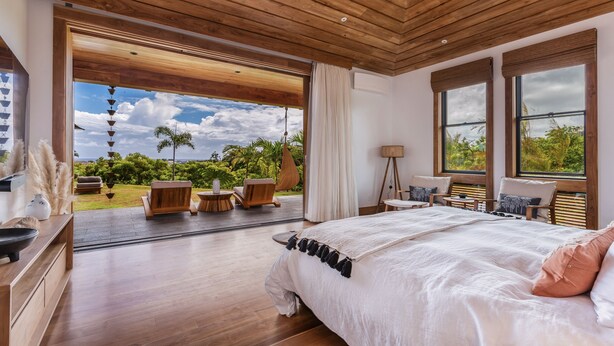 A spacious bedroom with a large bed, wooden ceiling, and a view of a lush garden.