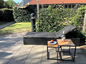 Un espace extérieur aménagé avec un canapé en osier et une petite table, une bouteille de vin et une assiette de fruits.